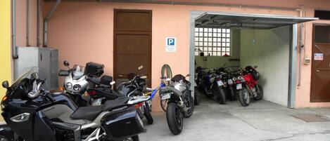 Un área de estacionamiento con varias motocicletas y bicicletas, la entrada de un edificio y un letrero de estacionamiento.