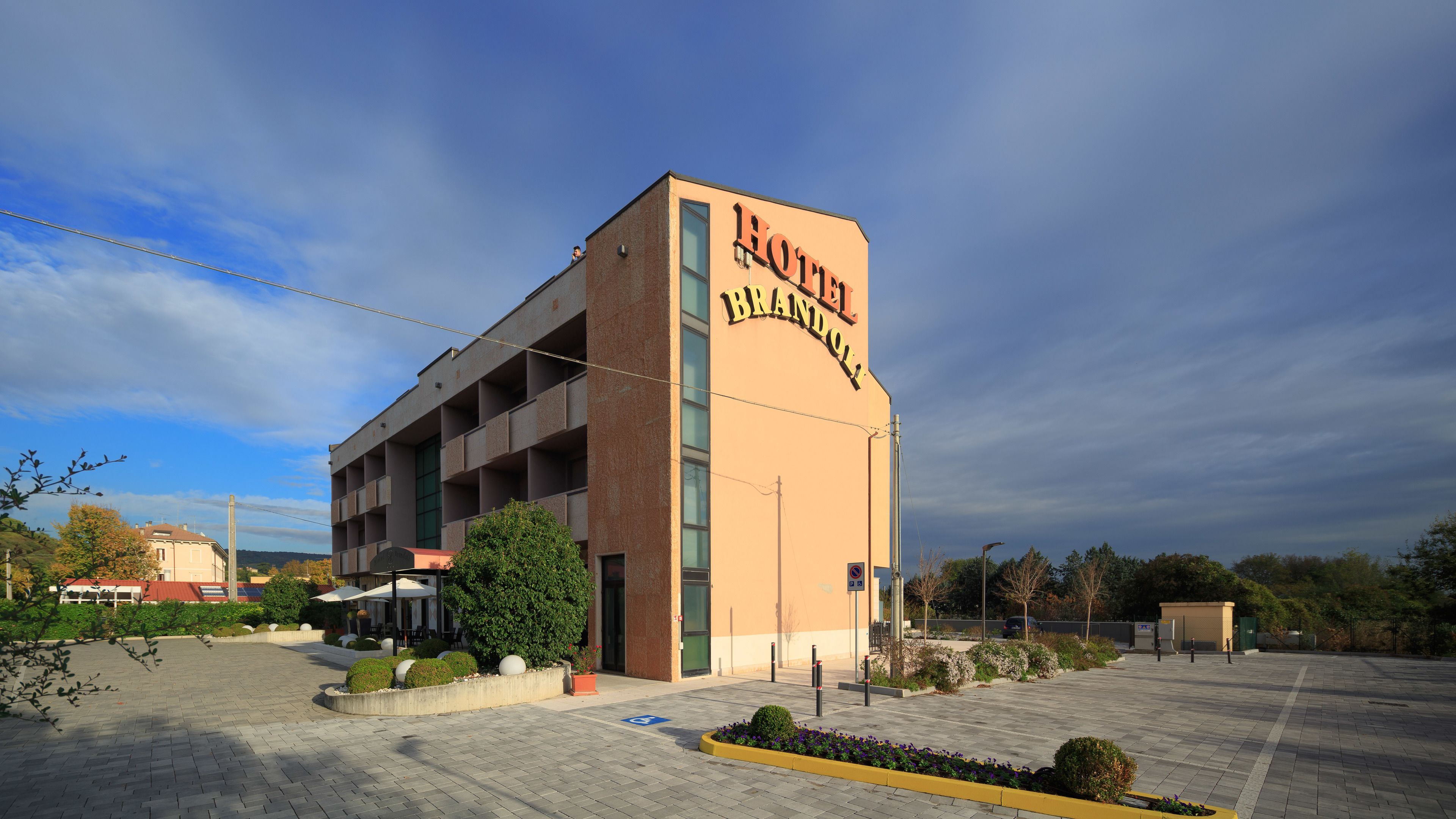 Un hôtel appelé «Hotel Brandol», doté d’une architecture moderne, se distingue par son entrée imposante et son parking, avec un ciel dégagé en arrière-plan.