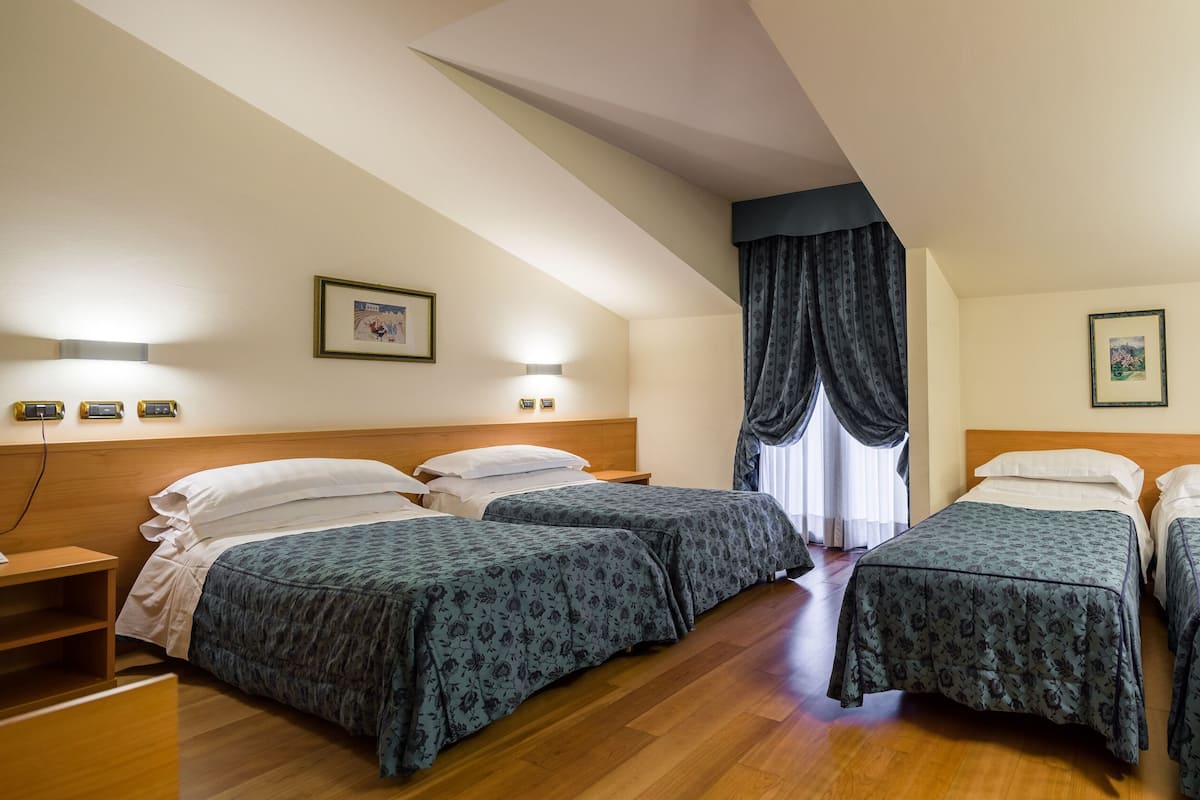 A hotel room with two single beds, wooden headboards, a desk with a chair, a nightstand, and a window with curtains.