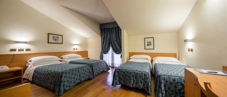 A hotel room with two single beds, wooden headboards, a desk with a chair, a nightstand, and a window with curtains.