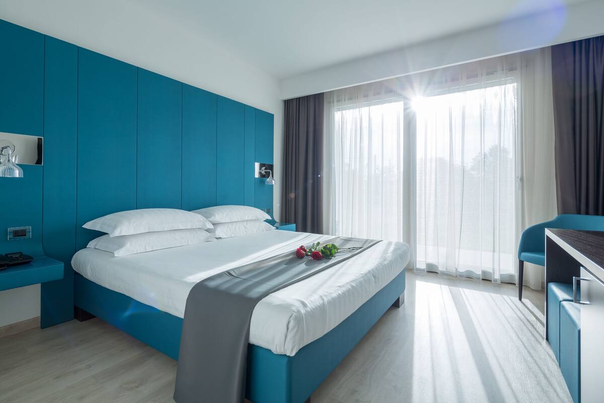 A modern hotel room with a large bed, a desk, and a view of the city through sheer curtains.