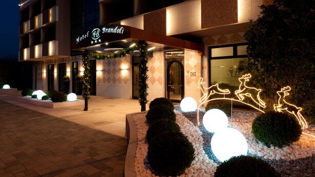 Entrada de un hotel con un cartel que dice 'Hotel Brandioli', rodeada de luces decorativas y jardines bien cuidados.
