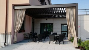 Un patio exterior con mesas y sillas, una pérgola y la fachada de un edificio con revestimiento de azulejos.
