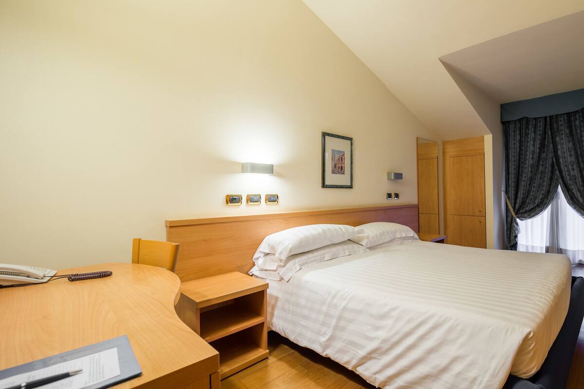 A hotel room with a bed, bedside table, lamp, and a desk with a chair.