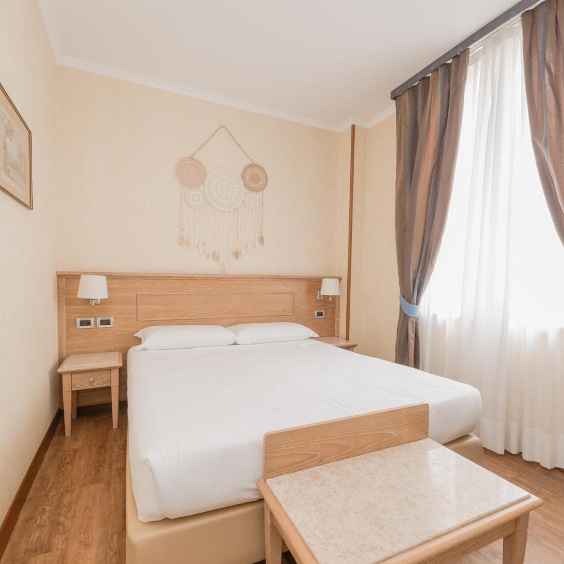 Hotellihuone, jossa on sänky, yöpöydät, seinäkoriste ja ikkuna, jossa on verhot.