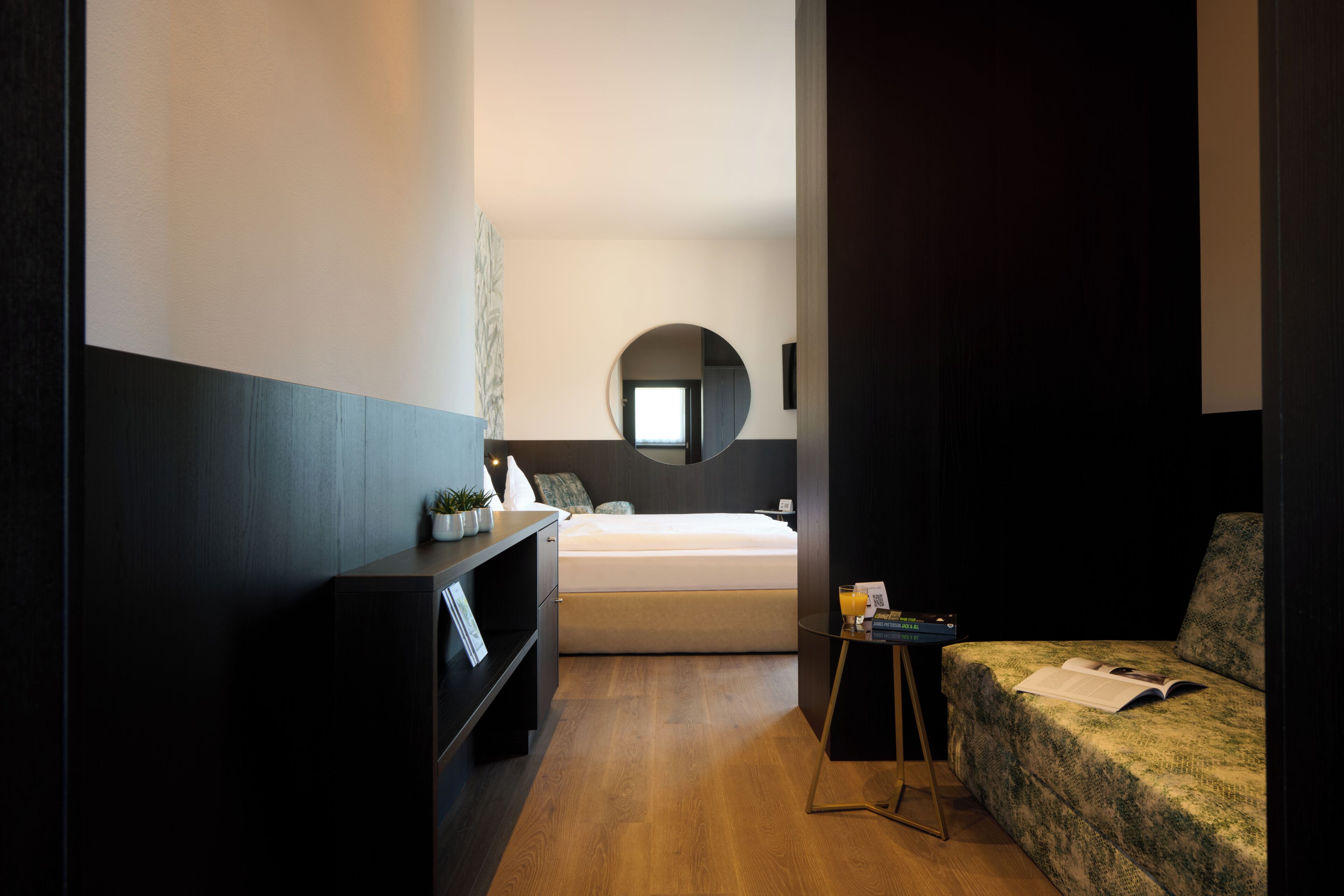 A modern hotel room with a bed, a small table with a book, a sofa, a mirror, and a shelf with decorative items.