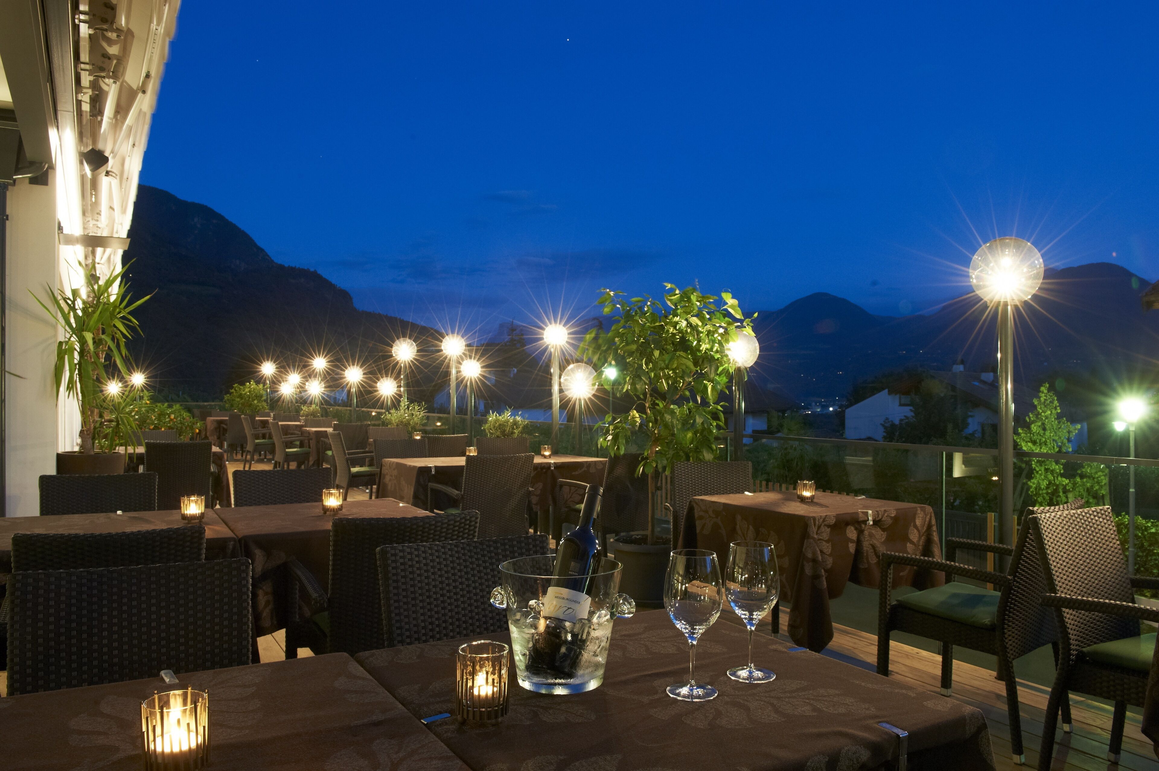 Un área de comedor al aire libre con mesas dispuestas para dos, un botellón de champán en un cubo de hielo, velas encendidas y vistas a montañas.
