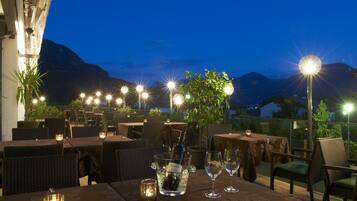 Un área de comedor al aire libre con mesas dispuestas para dos, un botellón de champán en un cubo de hielo, velas encendidas y vistas a montañas.