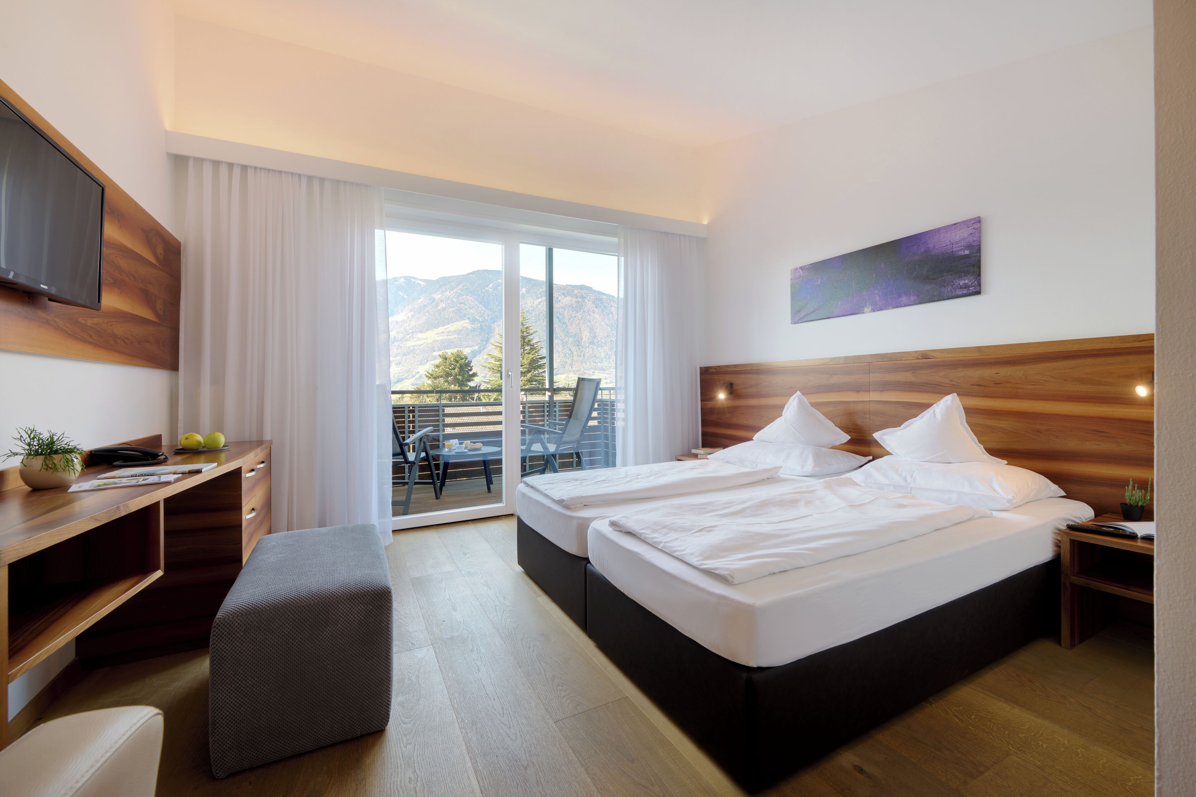 Habitación de hotel con cama, escritorio, silla, televisor y balcón con vistas a las montañas.