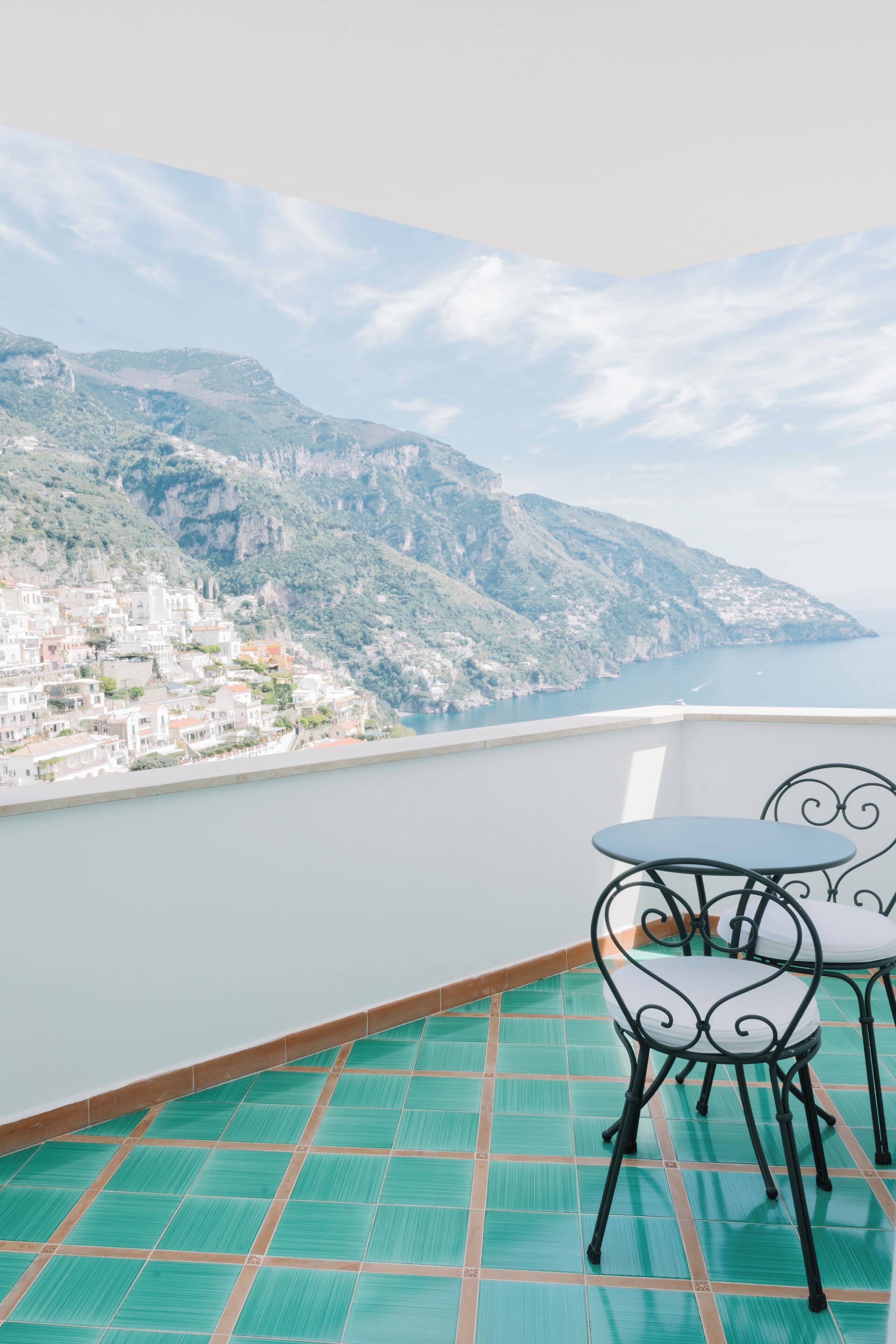 A balcony with a view of a coastal town, a tiled floor, and two metal chairs.