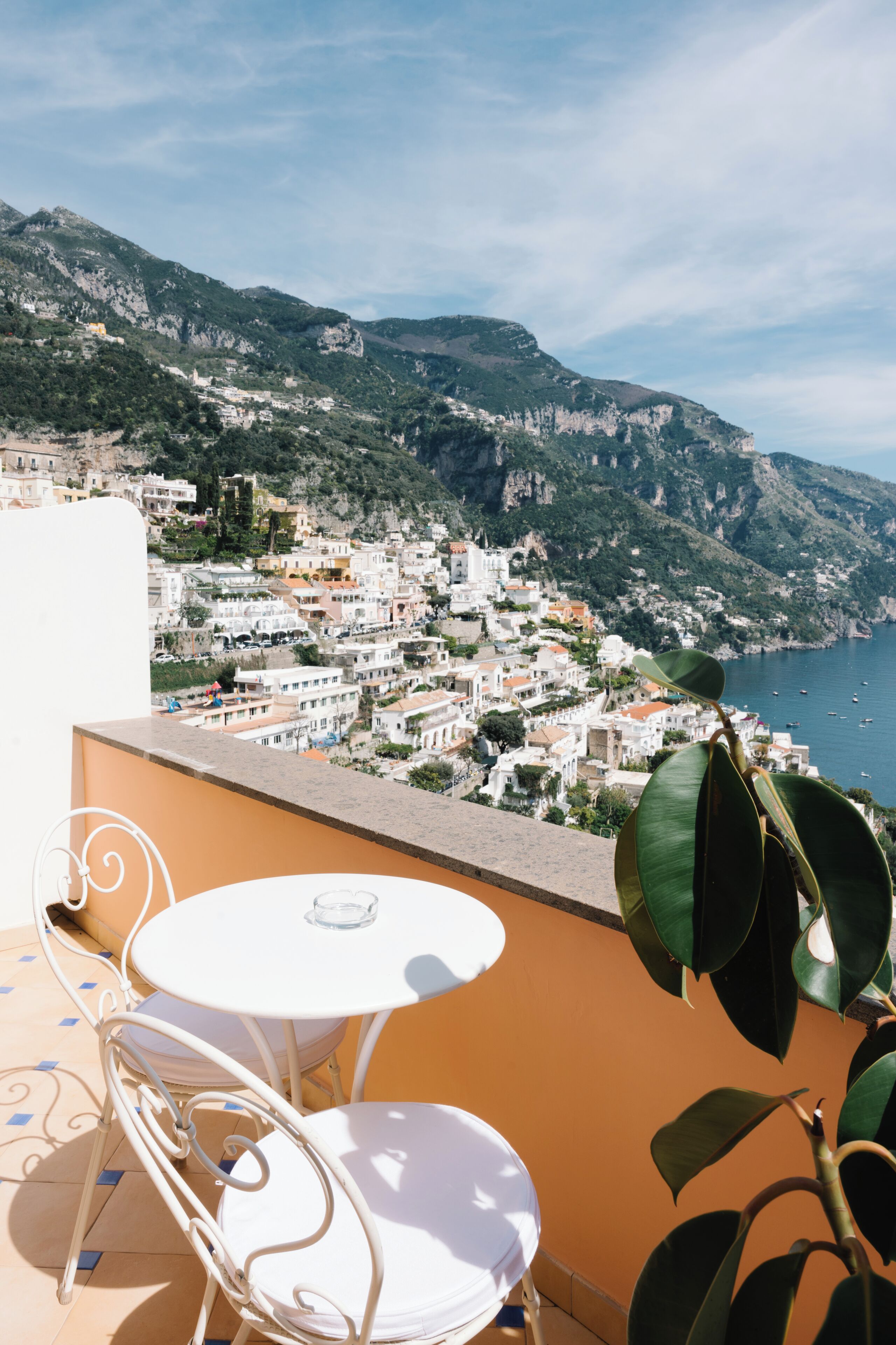 A terrace with a white table and chair, overlooking a coastal town with buildings and cliffs.