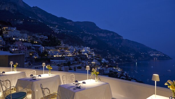 Une terrasse sur le toit, avec des tables dressées pour les repas, offrant une vue sur une ville côtière et la mer.