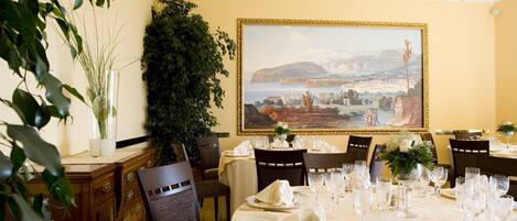 A dining area with a table set for six, a large framed painting on the wall, and a potted plant on the side.