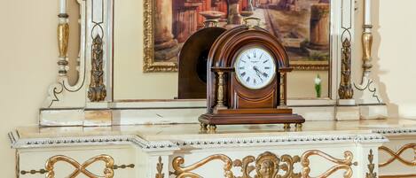 Un reloj de pared con diseño clásico, situado sobre una mesa con cubierta de mármol y detalles ornamentales blancos y dorados.