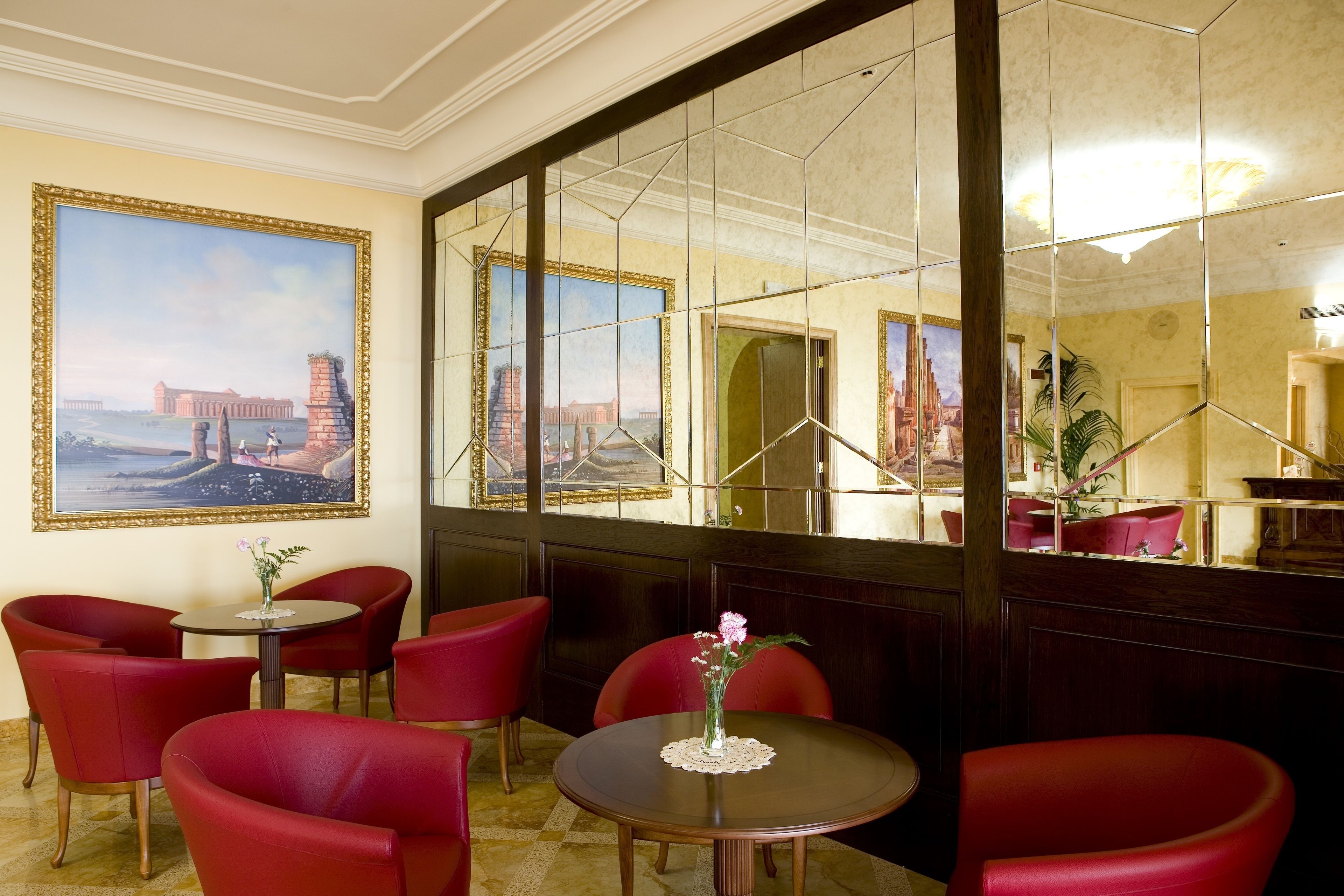 A hotel lobby with red chairs, a round table with a vase of flowers, and a large mirror reflecting the room.