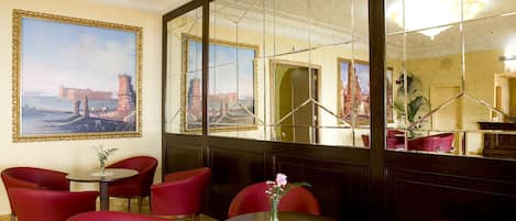 A hotel lobby with red chairs, a round table with a vase of flowers, and a large mirror reflecting the room.