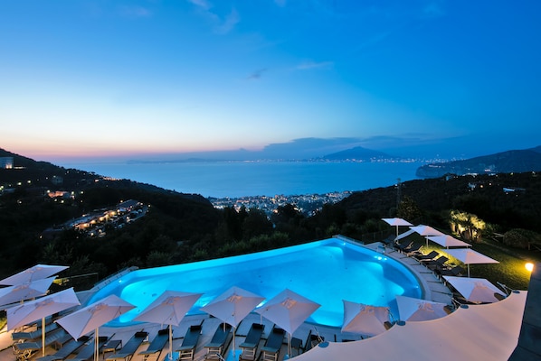 A pool area with lounge chairs and umbrellas, overlooking a coastal cityscape at dusk.