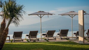 Un espace aménagé au bord de la piscine, avec des chaises longues et des parasols, un palmier, et une vue sur une étendue d’eau, avec des montagnes en arrière-plan.