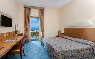 Habitación de hotel con cama, escritorio, silla, televisión y balcón con vistas.