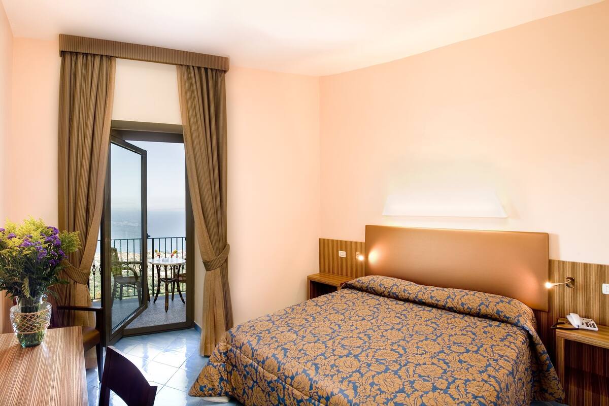 A hotel room with a bed, a TV, a desk with a vase of flowers, a balcony with a table and chairs, and a view of the sea.