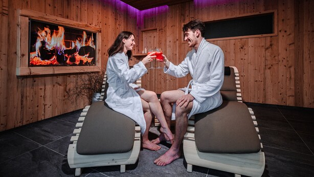 Two people in white robes seated on cushioned chairs in a wooden room with a fireplace and a television displaying a fire scene. - Albergo Moderno (Fuipiano Valle Imagna)