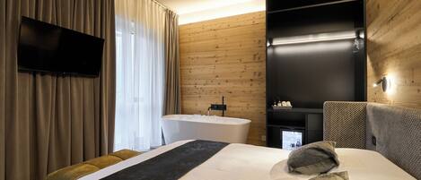 A modern hotel room with a large bed, a flat-screen TV mounted on the wall, a built-in bathtub, and a wooden accent wall.