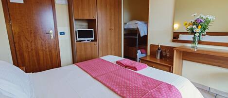 Habitación de hotel con una cama grande, un armario de madera, un escritorio con espejo y un pequeño estante con un jarrón de flores.