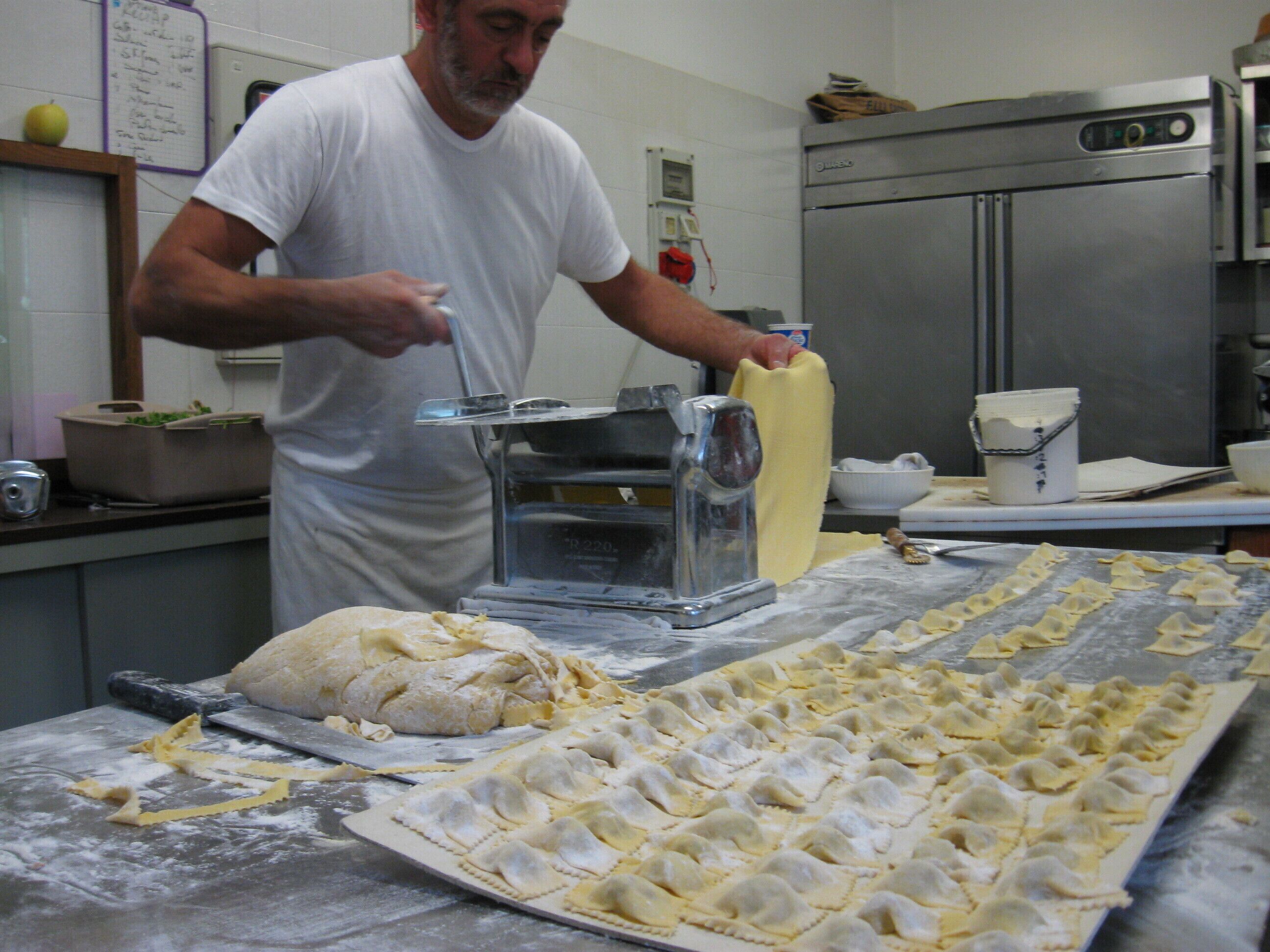 A kitchen with a large table covered in flour, a man preparing dough, and various kitchen appliances.