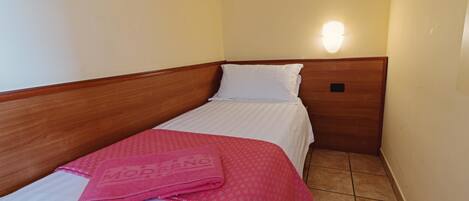 A small, single-bed room with a wooden headboard, a white bedspread, a pink towel, and a wall-mounted light.
