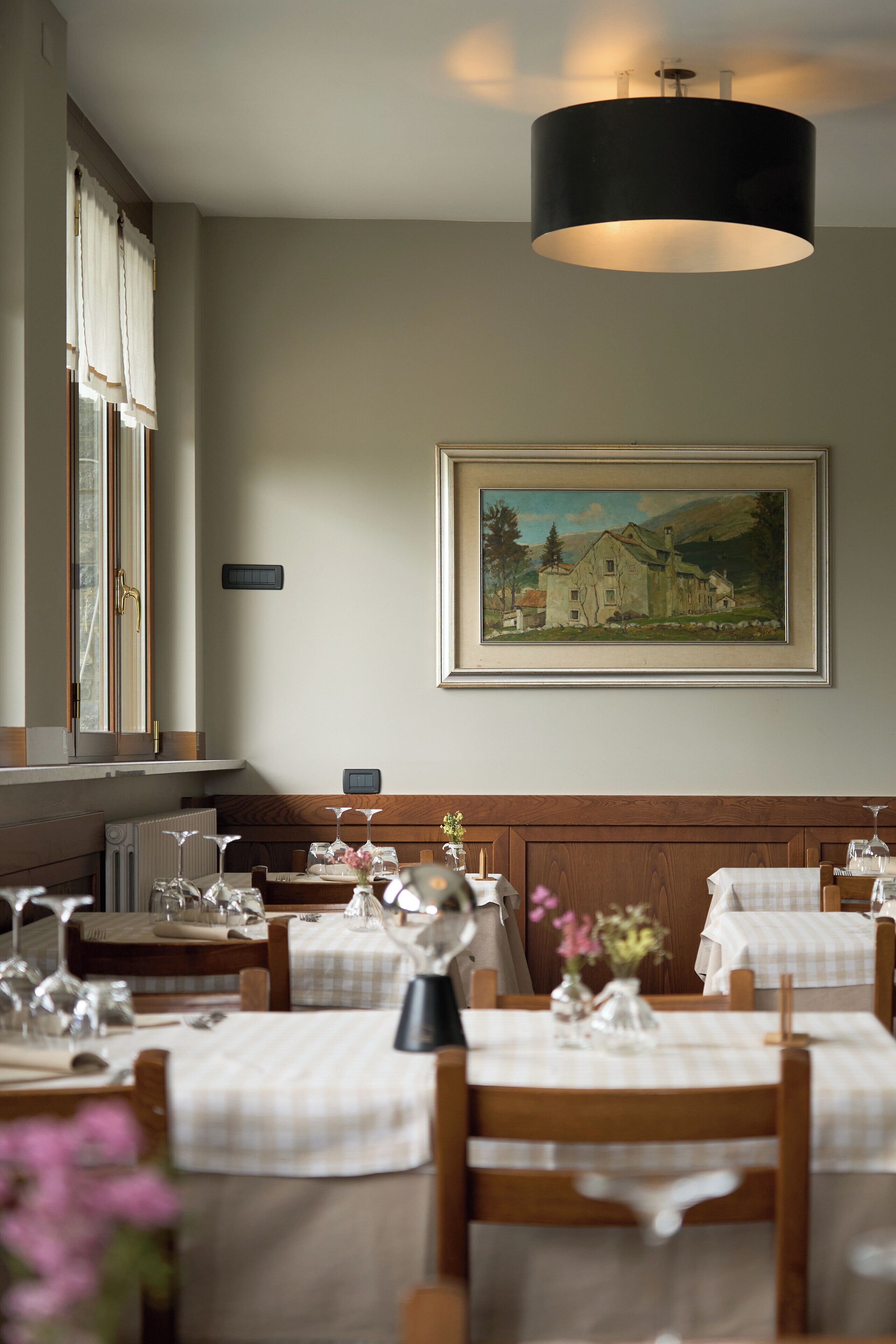 A dining area with tables set for a meal, a painting on the wall, and a window with curtains.