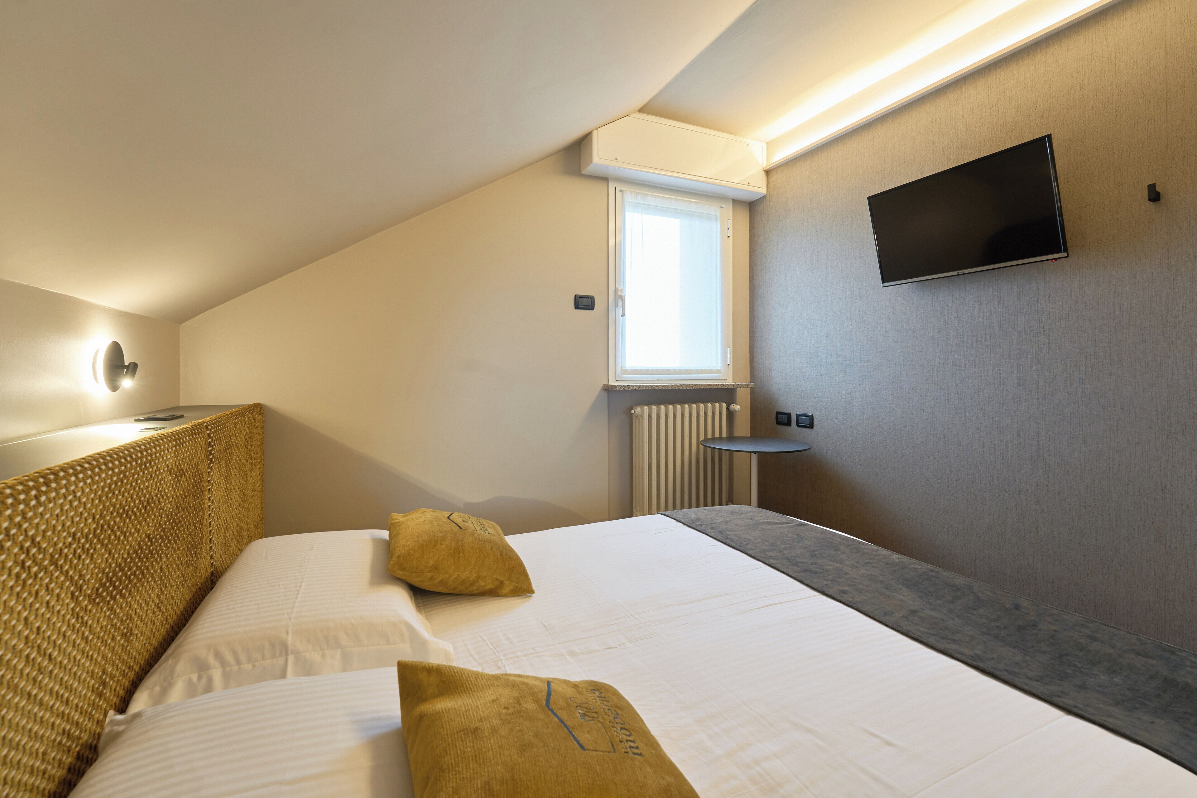 Habitación de hotel con cama, dos almohadas, mesita de noche, televisor montado en la pared y ventana con aire acondicionado.