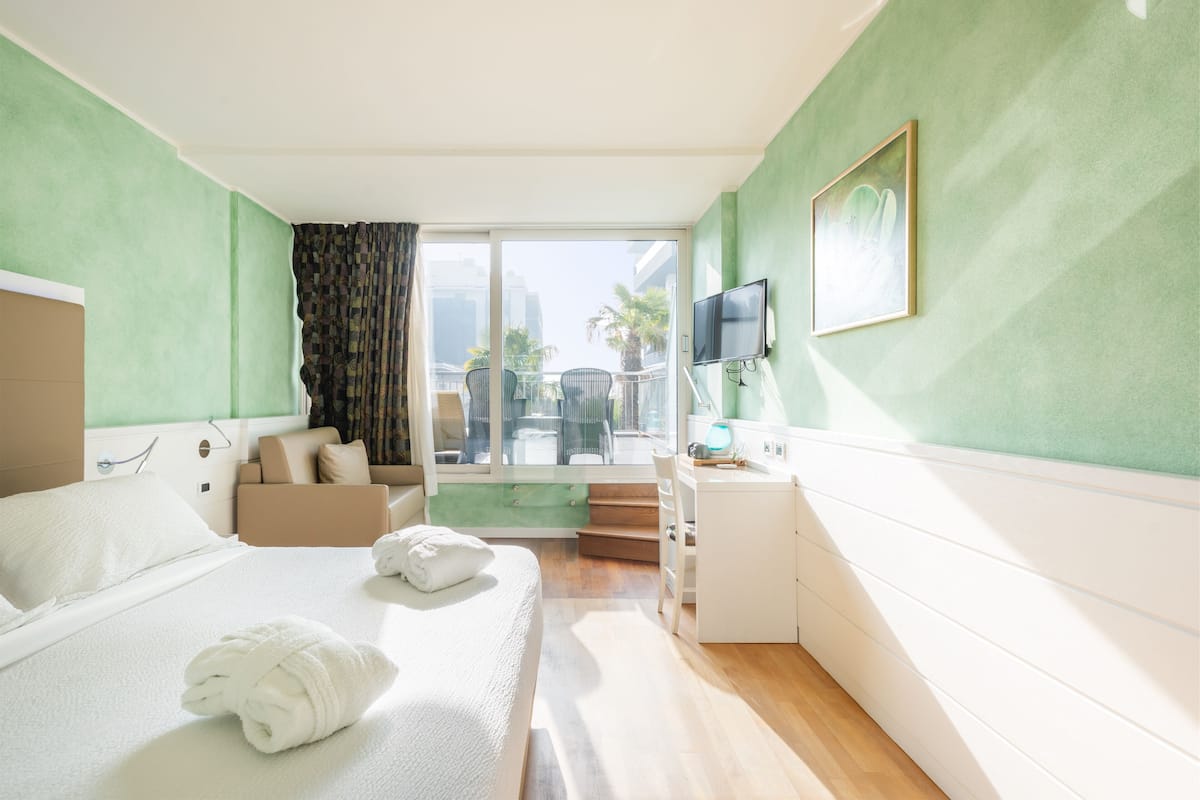 A hotel room with a bed, a desk, a TV, and a view of palm trees and buildings.