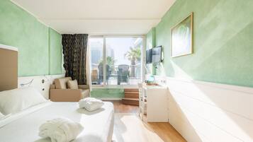A hotel room with a bed, a desk, a TV, and a view of palm trees and buildings.