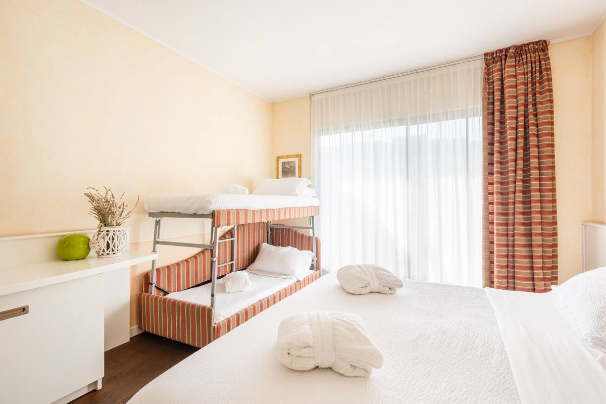 A hotel room with bunk beds, a bedside table with a vase, and a large bed with towels.