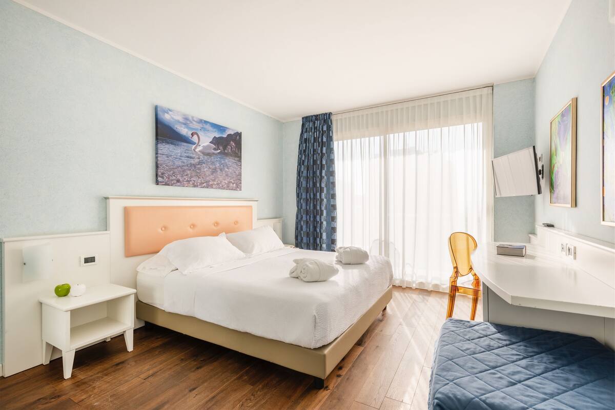 A hotel room with a bed, a desk, a chair, a nightstand, and a painting on the wall.