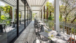 Un espacio de comedor al aire libre con mesas y sillas, rodeado de follaje y una barandilla blanca.