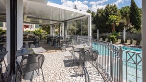 Un patio al aire libre con mesas y sillas, una piscina y vistas a árboles y edificios al fondo.