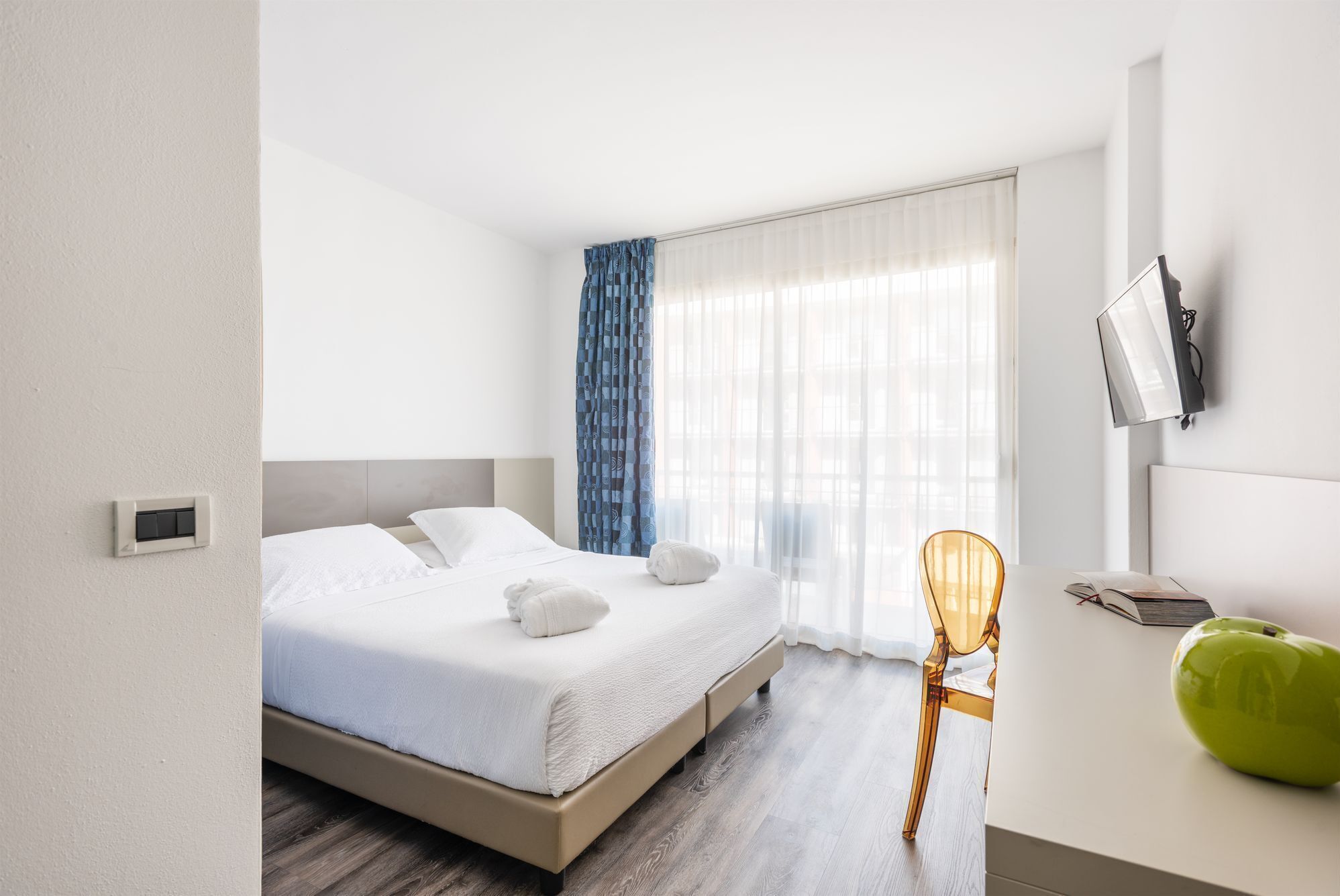 Una habitación de hotel moderna con una cama, un escritorio con una silla, un televisor y un ventanal con cortinas.