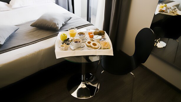 A neatly made bed with white linens and pillows, a breakfast tray with various foods and drinks, and a modern chair with a reflective surface. - Hotel Ilga (Collecchio)