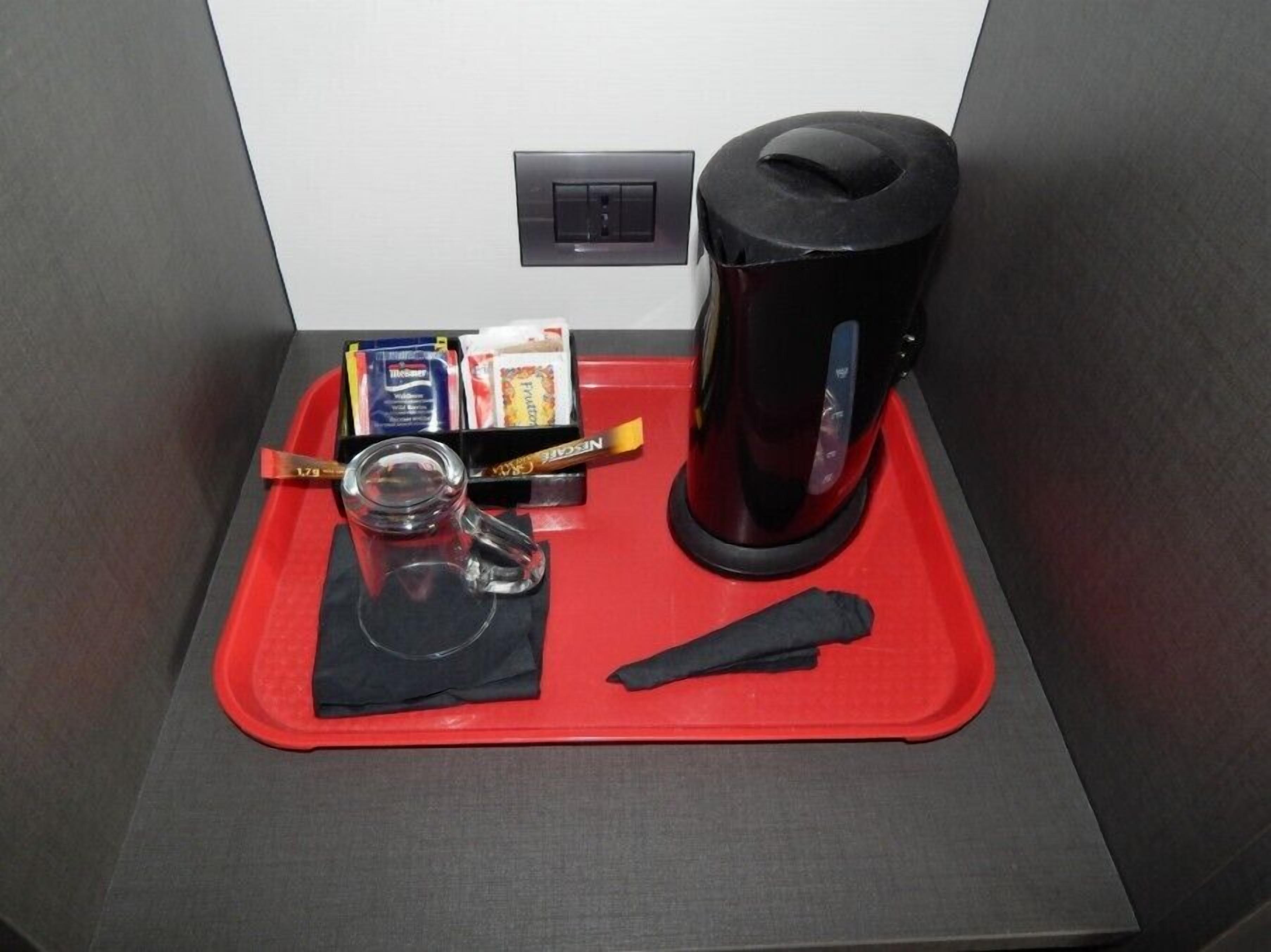 A small kitchenette area with a coffee maker, a glass, a spoon, a packet of sugar, and a box of tissues on a red tray.
