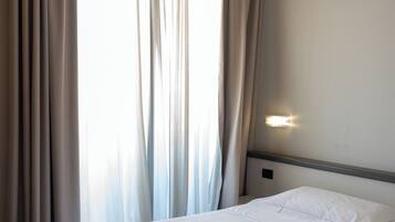 A hotel room with a large bed, beige curtains, a wall-mounted light, and a wooden floor.