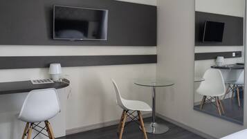 A modern hotel room with a flat-screen TV mounted on the wall, a small round table with a chair, a desk with a chair, and a large mirror reflecting the room.