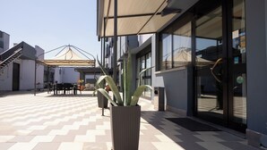 An outdoor area with a patterned floor, a potted plant, outdoor furniture, and a building entrance with glass doors. - Hotel Ilga (Collecchio)