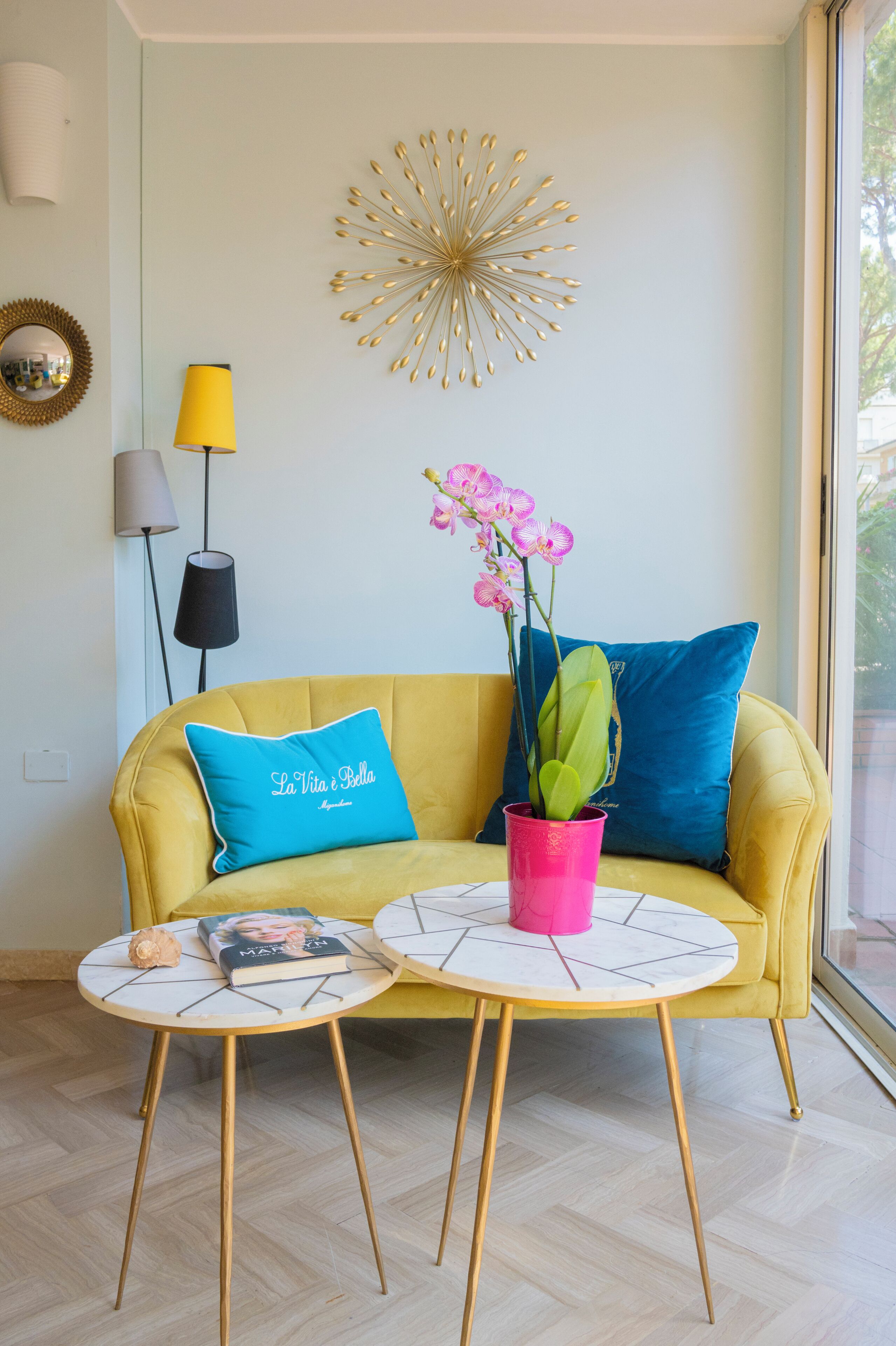 A yellow sofa with a blue pillow and a pink vase with flowers. There is a gold decorative piece on the wall and a lamp with a black shade.