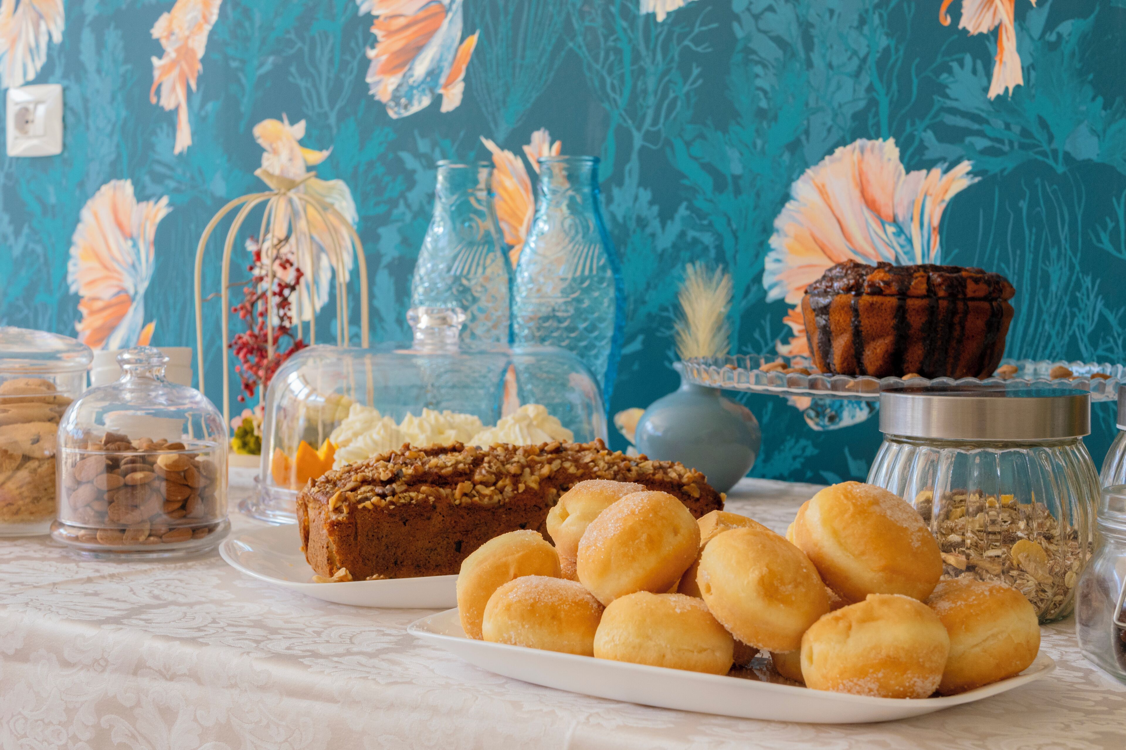 A table with various pastries and desserts, including a cake and doughnuts, with decorative glass containers and a floral wallpaper background.
