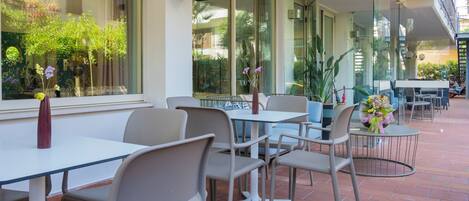 Un área de estar al aire libre con mesas y sillas, ventanas amplias con vistas a la vegetación y un diseño moderno.