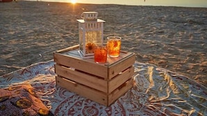 A wooden crate on a blanket with a lantern and drinks on a beach at sunset.