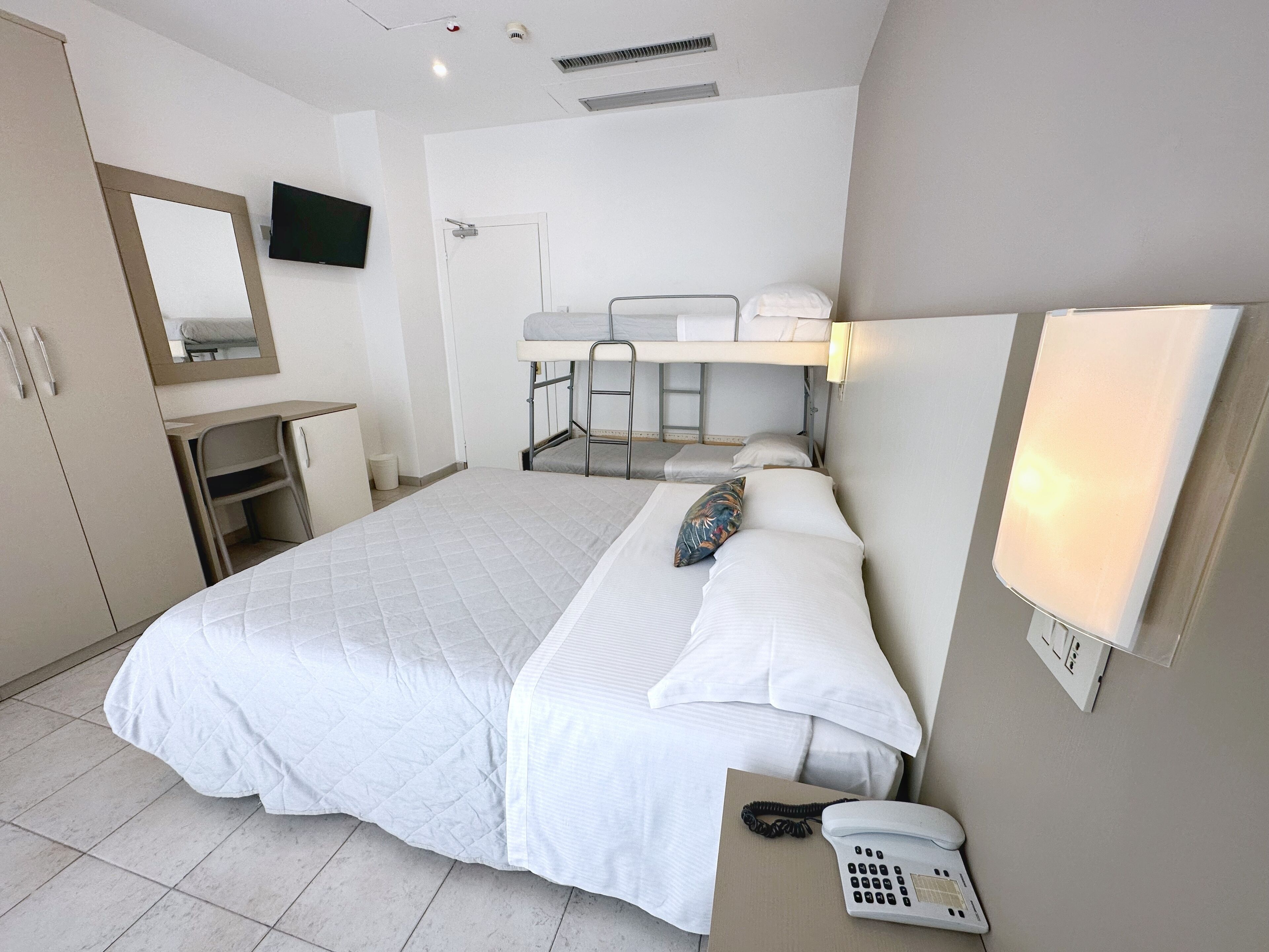 Habitación de hotel con una cama grande, un escritorio, una silla, un espejo, un televisor, una cama litera y un teléfono.