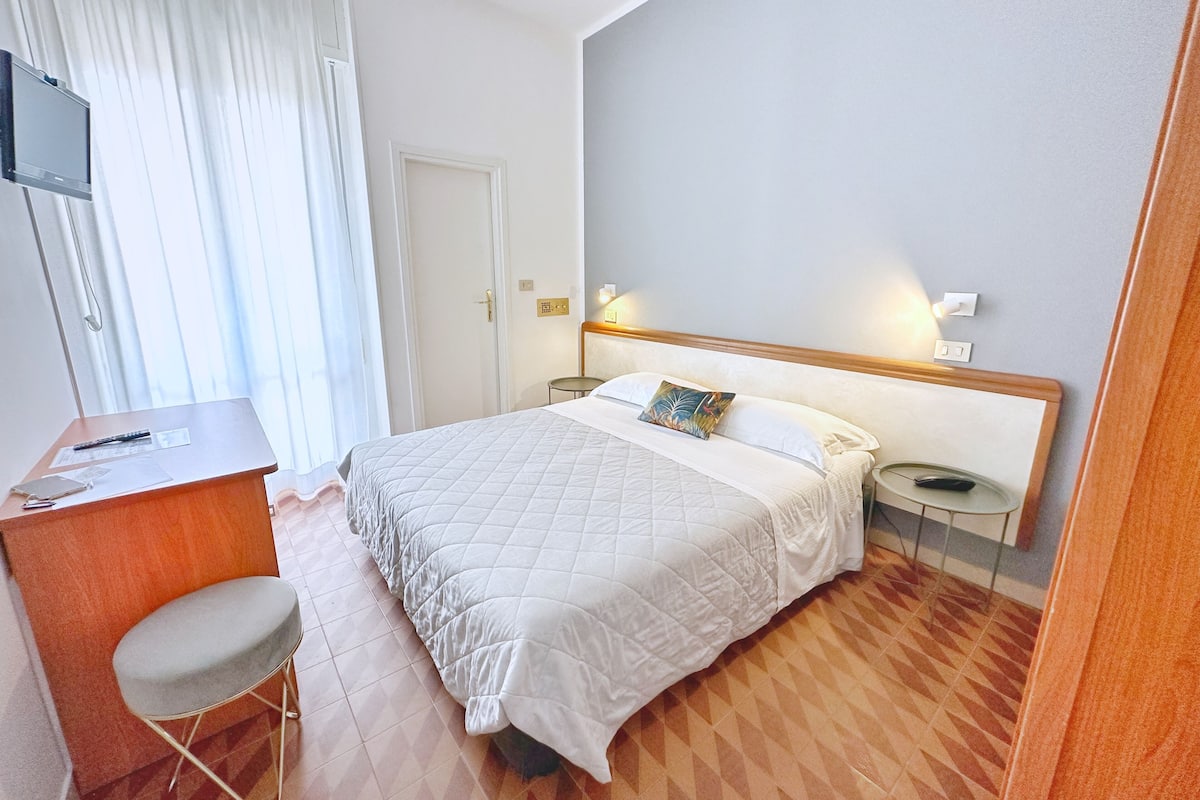 A hotel room with a bed, a desk, a chair, a TV, and a nightstand with a lamp.