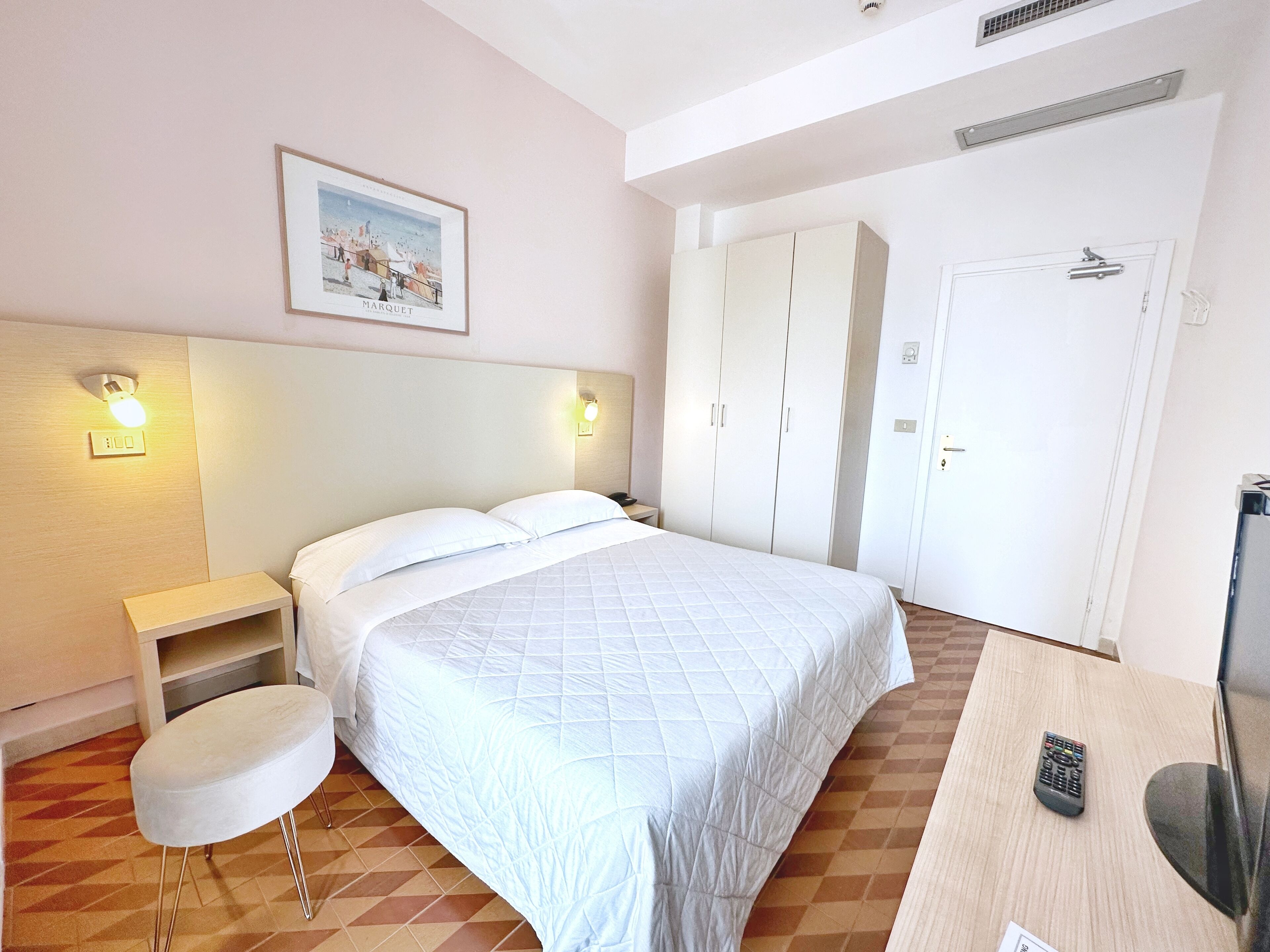 A hotel room with a bed, a desk with a TV, a chair, a wardrobe, and a picture on the wall.